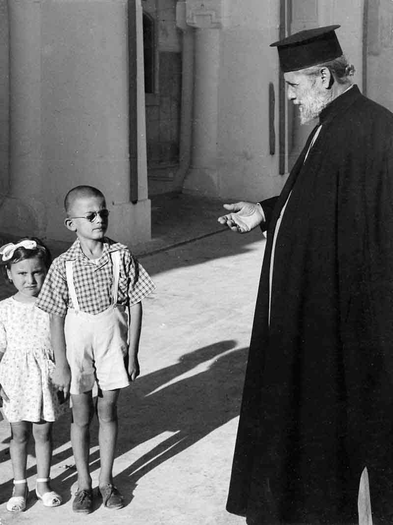 An old black and white photo depicting a greek priest standing with two children, capturing a moment of innocence and faith.