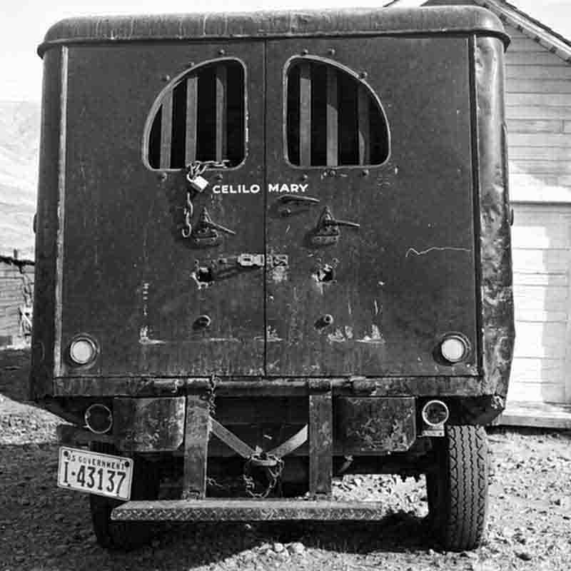 A black and white photograph by Peter Bock-Schroeder from 1956, showing the rear of a dark, weathered transport van parked on uneven dirt ground. The double back doors feature two small, arched windows with thick vertical metal bars, secured with a heavy chain and padlock. The name 'CELILO MARY' is hand-painted in white capital letters across the center of the doors. A 'U.S. GOVERNMENT' license plate numbered 'I-43137' is visible on the lower left.