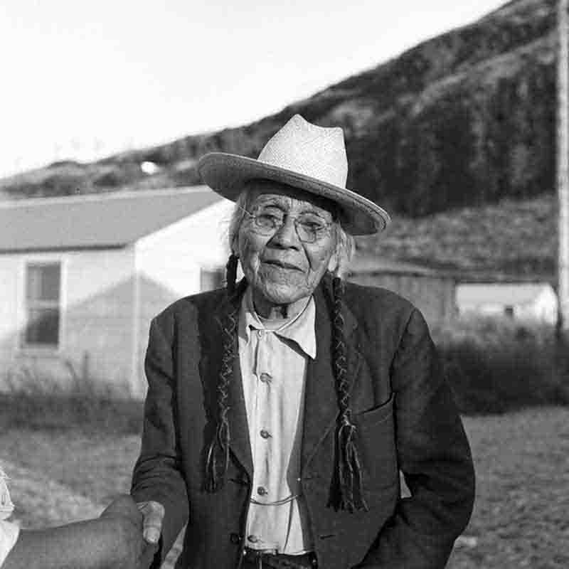 A black and white portrait by Peter Bock-Schroeder from 1956, featuring Chief Tommy Thompson of the Wyam tribe at Celilo Falls. The Chief is shown from the chest up, wearing a light-colored hat, eyeglasses, and a dark jacket over a button-down shirt. His hair is styled in two long, dark braids that hang over his shoulders.