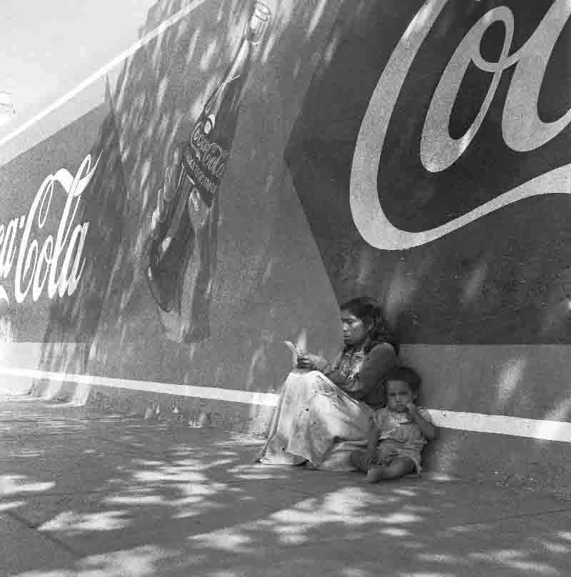 Peruvian native mother with toddler sitting on the street in Lima in front of a Coca Cola advertisement