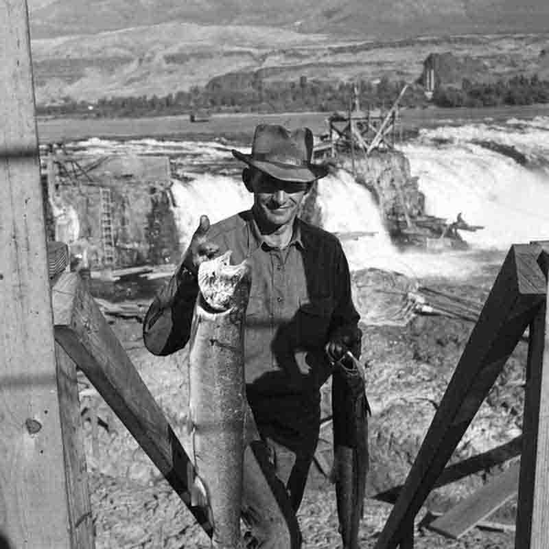 A black and white medium shot by Peter Bock-Schroeder, taken in 1956, featuring a smiling fisherman at Celilo Falls. The man, wearing a wide-brimmed fedora and a button-down shirt, stands on a wooden platform holding a large, freshly caught salmon by its gills.