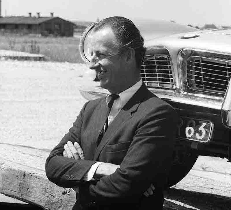 A well-dressed man, Graf Goertz,  standing in front of a BMW 507 car, exuding professionalism and elegance