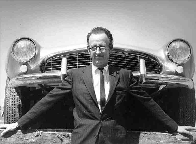 A sophisticated man, clad in a tailored suit, exudes opulence while standing alongside a magnificent BMW 507 sports car.