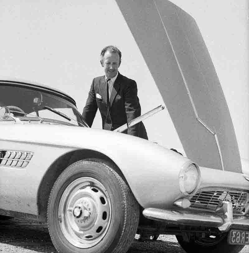 A well-dressed man, Graf Goertz,stands on a BMW 507 sports car with the hood open.