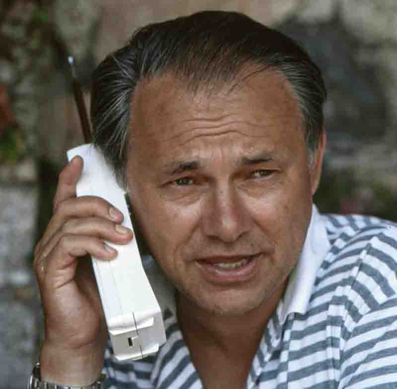 A close-up photograph by Ingeborg Bock-Schroeder of Hubert Burda with dark, slightly graying hair and a striped shirt, talking on a large, white mobile phone with an antenna.