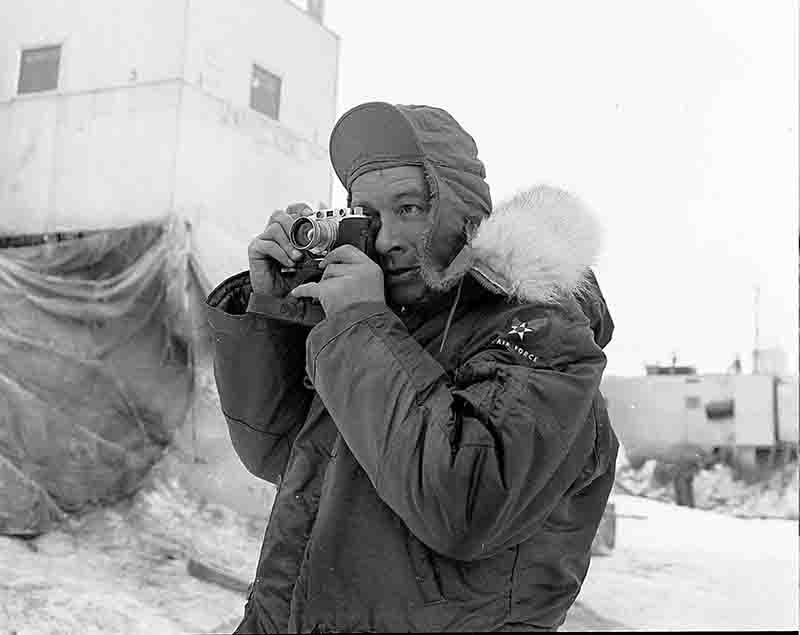 A black and white photograph of German photo reporter Peter Bock-Schroeder captured in a frigid, outdoor environment. He is dressed for extreme cold, wearing a heavy, dark US Air Force parka. The camera Peter Bock-Schroeder is using in this photograph is a Leica rangefinder, M3 model.
