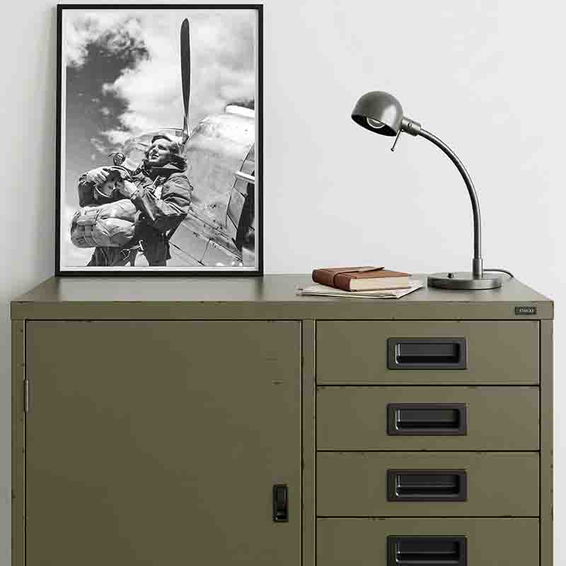 A framed, black and white photograph of pilot standing next to the nose and propeller of an aircraft. The framed photo is displayed on a green metal cabinet next to desk lamp and a stack of notebooks.