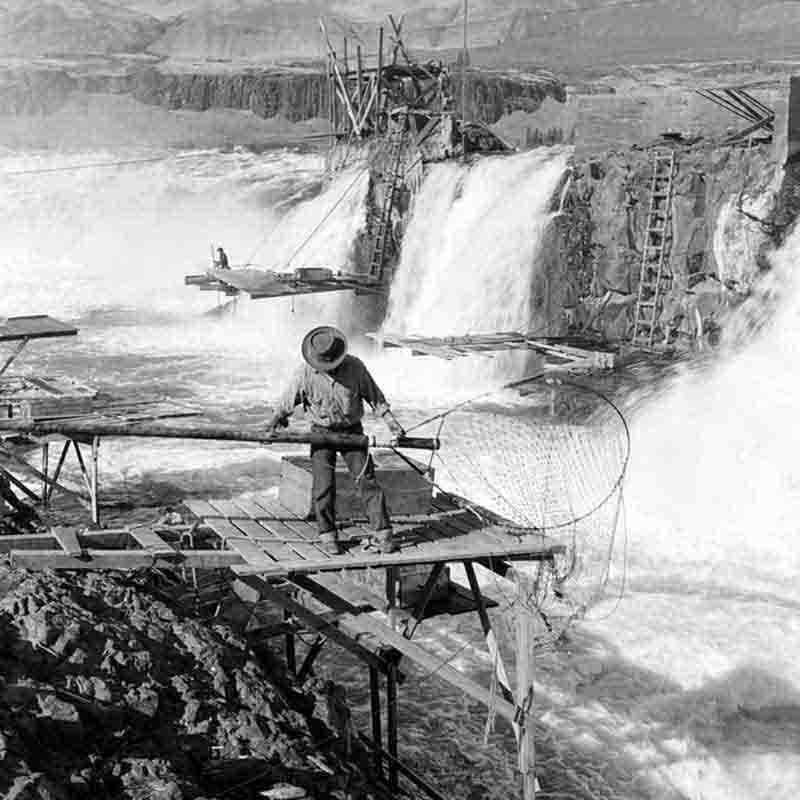 A black and white photograph by Peter Bock-Schroeder from 1956, capturing a fisherman standing on a precarious wooden platform cantilevered over the churning white water of Celilo Falls.