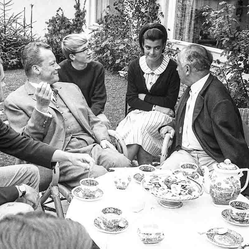Willy Brandt, Chancellor of West Germany, sitting in a garden surrounded by green plants, deep in conversation. The scene is peaceful and intimate, showcasing Brandt's approachable and down-to-earth nature.